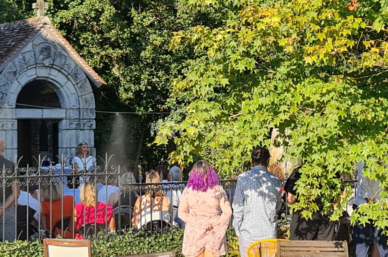 Un groupe de personnes assiste à un événement en plein air près d'une petite chapelle en pierre entourée de verdure.