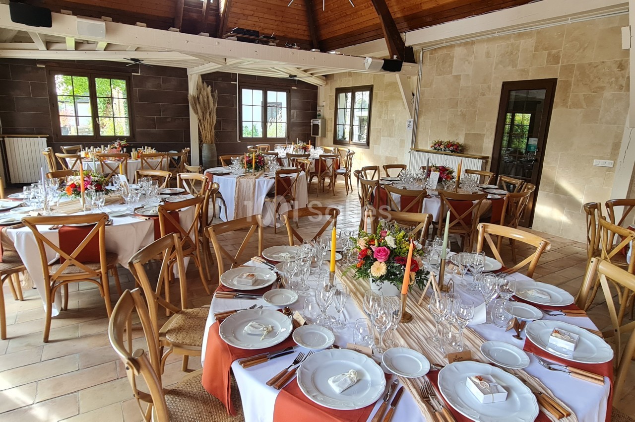 Salle de réception lumineuse avec tables rondes décorées de nappes blanches, chemins de table orange et arrangements floraux.