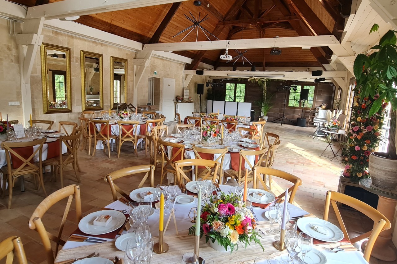 Salle de réception lumineuse avec tables dressées, décorations florales colorées et poutres apparentes en bois.