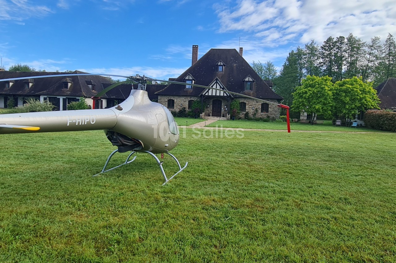 Hélicoptère posé sur une pelouse devant un grand bâtiment en pierre entouré d'arbres et de verdure.