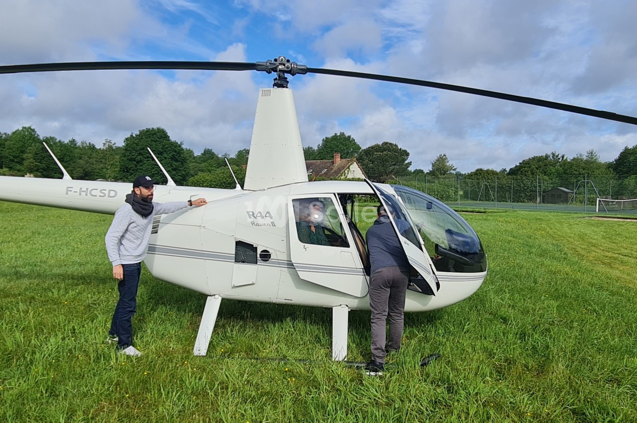 Deux hommes inspectent un petit hélicoptère blanc stationné sur une pelouse, entouré de verdure et de ciel nuageux.