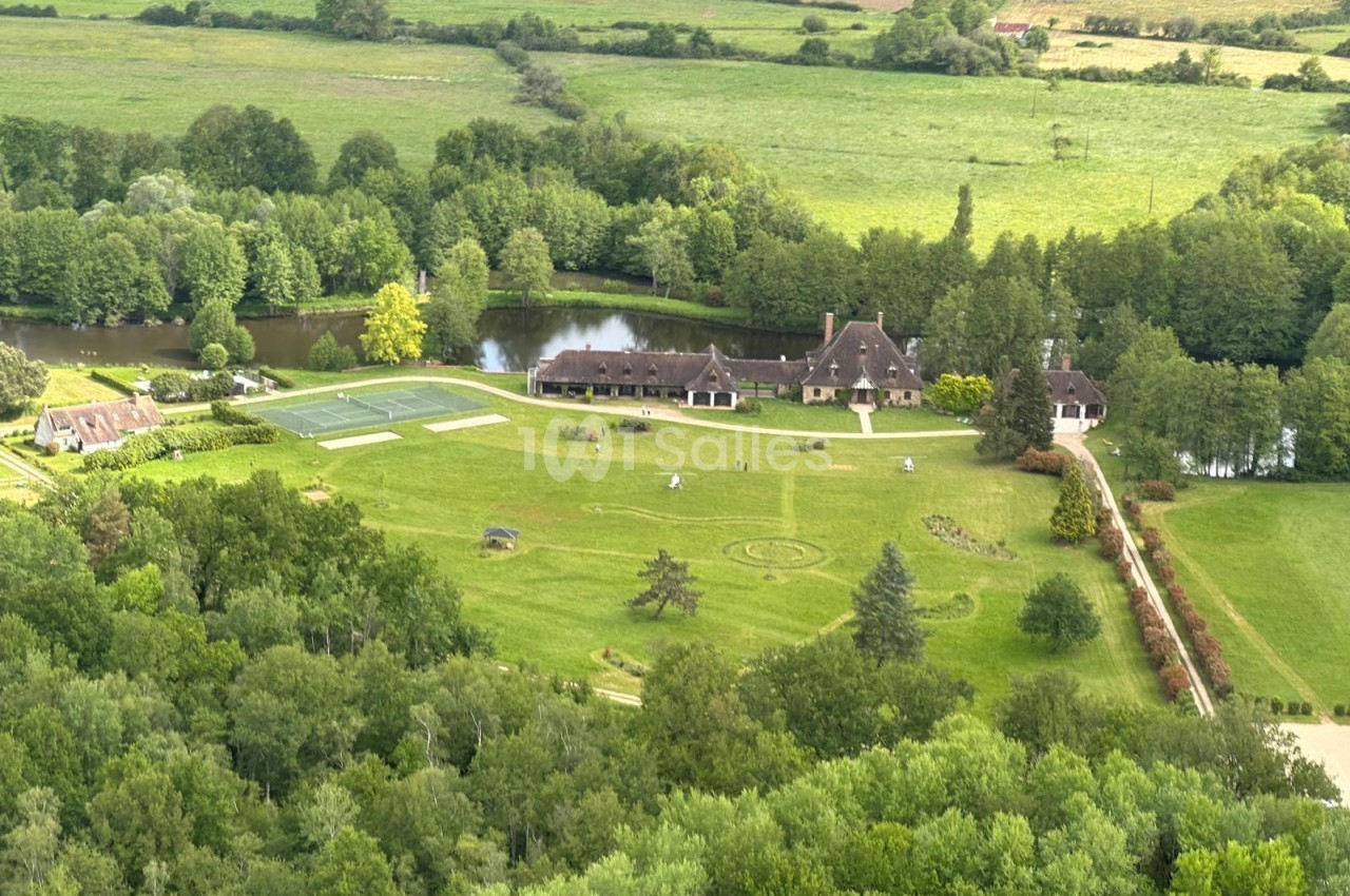 Vue aérienne d'un domaine avec bâtiments, pelouses aménagées, court de tennis et étang, entouré de verdure.