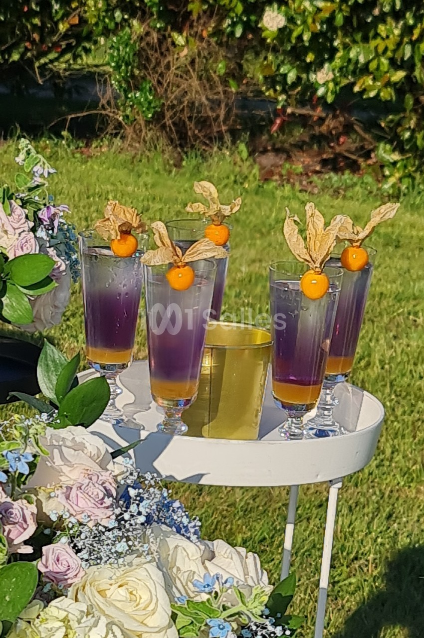 Verres de cocktail violets garnis de physalis, posés sur une table blanche décorée de fleurs, en extérieur.