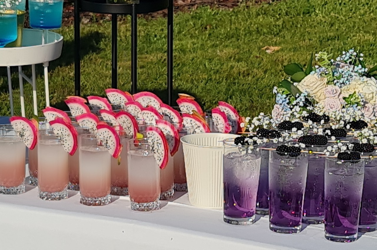 Table avec des boissons colorées, dont des cocktails roses à la pitaya, violets avec mûres et verts, décorée de fleurs.