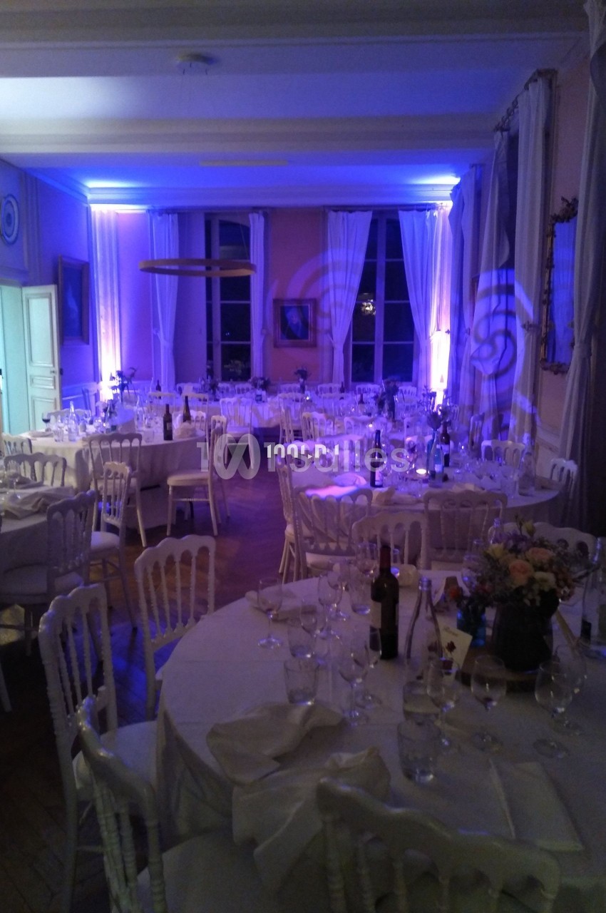 Salle de réception éclairée en bleu avec tables rondes dressées, chaises blanches et décorations élégantes.