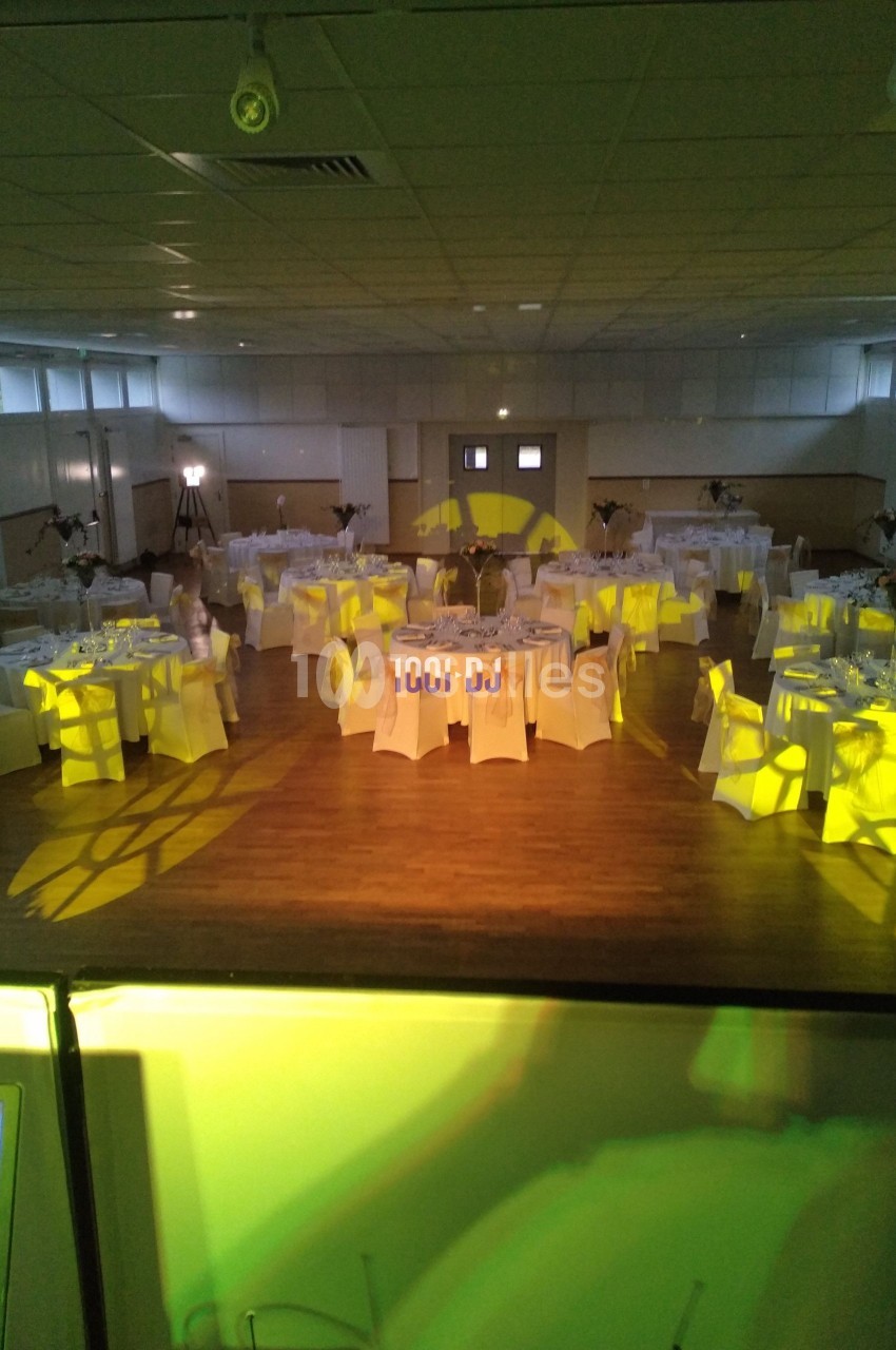 Salle de réception décorée avec des tables rondes dressées, éclairage jaune diffus et parquet en bois.