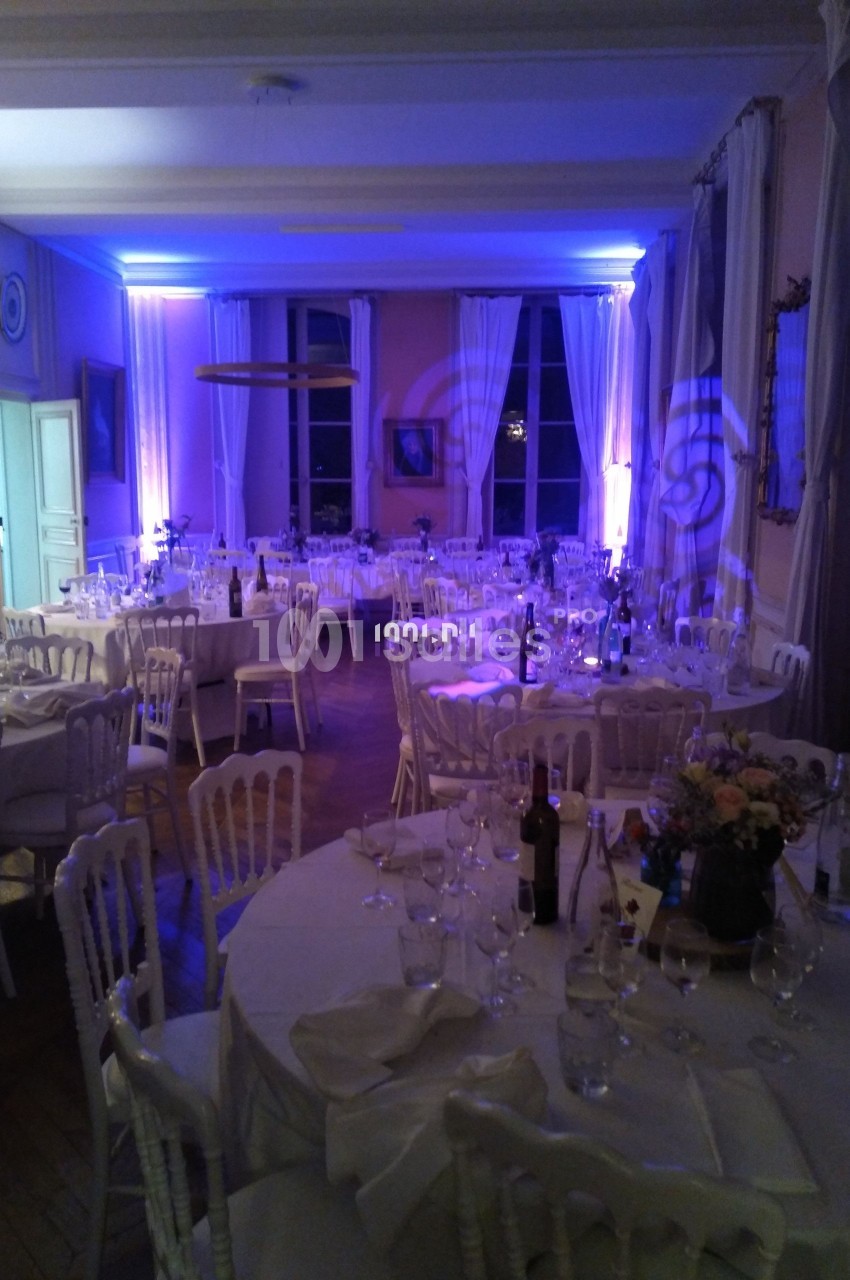 Salle de réception éclairée en bleu avec tables rondes dressées, chaises blanches et décorations élégantes.