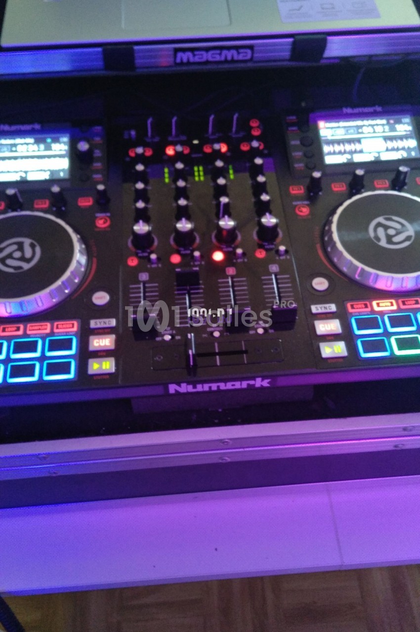Table de mixage DJ Numark avec deux platines, des boutons lumineux et un écran, posée sur un support.