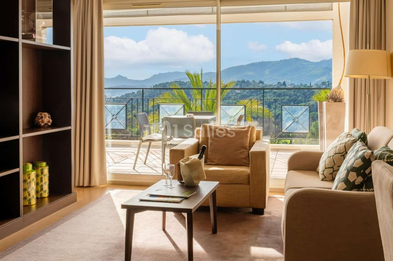 Salon lumineux avec canapé, fauteuil, table basse et vue sur une terrasse donnant sur des montagnes et un ciel dégagé.