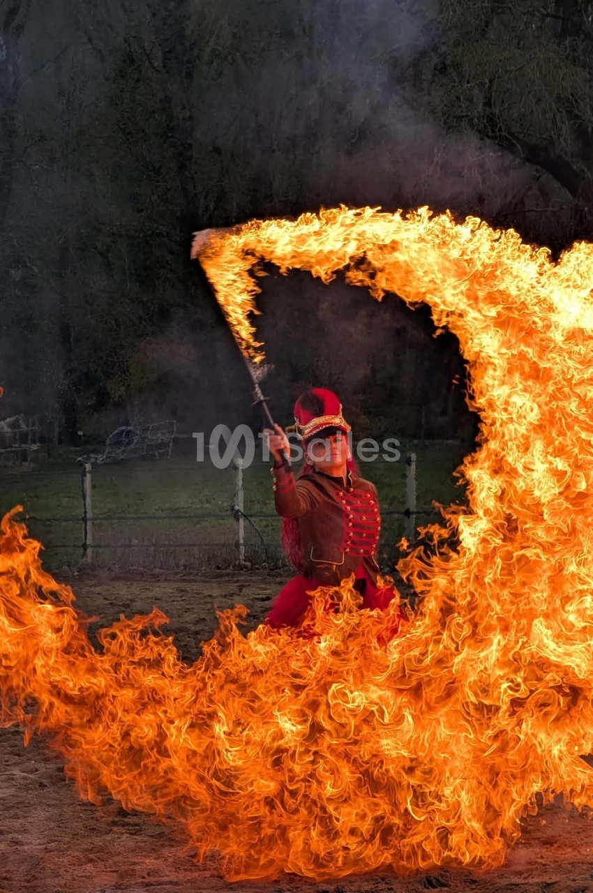 Une femme en costume rouge manipule un bâton enflammé, créant un arc de feu dans un espace extérieur.