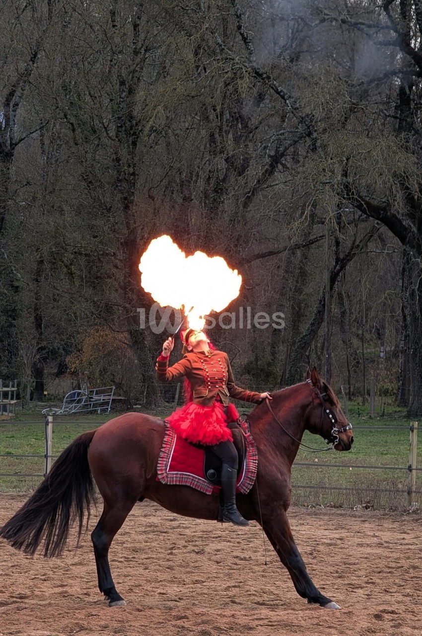 Une femme en costume rouge souffle du feu tout en étant assise sur un cheval dans une arène en plein air.