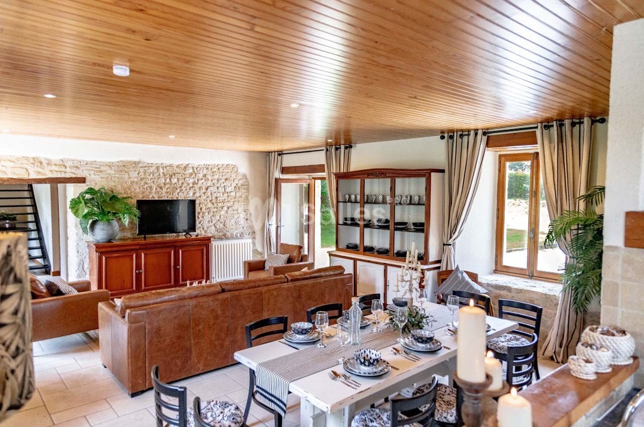 Salle à manger et salon lumineux avec mobilier en bois, canapé en cuir, murs en pierre et grandes fenêtres.