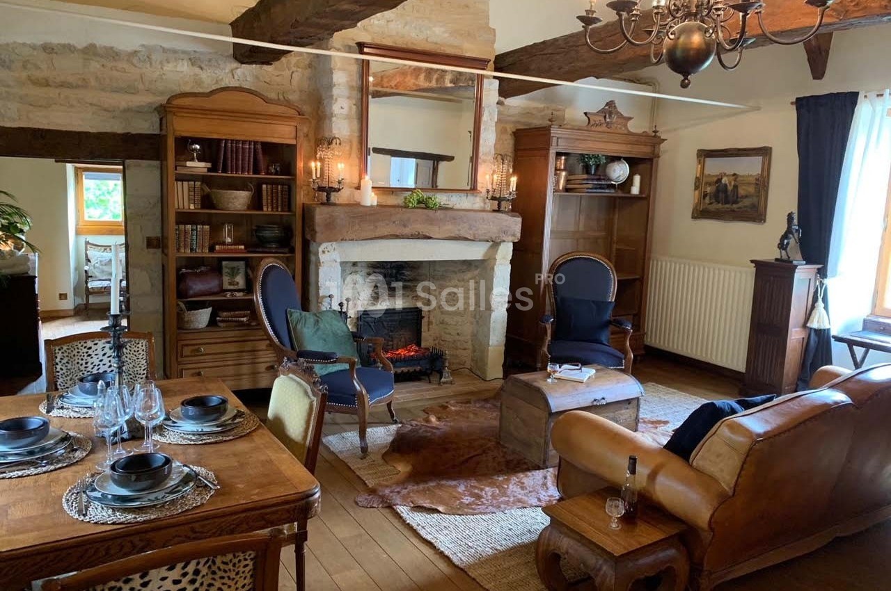Salon rustique avec poutres apparentes, cheminée en pierre, meubles en bois et coin repas avec table dressée.