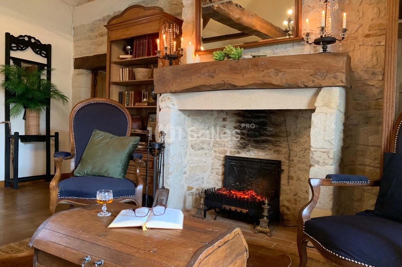 Cheminée en pierre allumée dans un salon rustique avec fauteuils, table basse en bois et étagères remplies de livres.