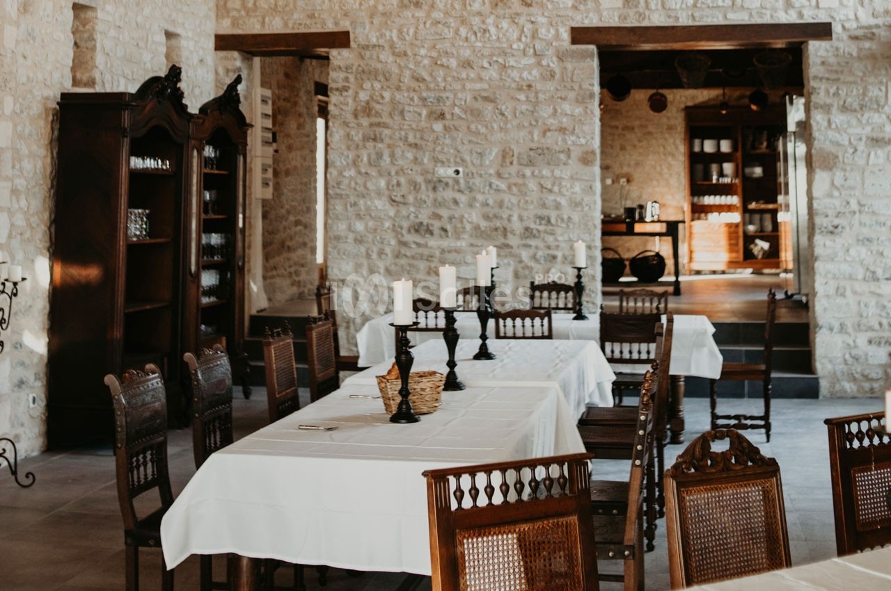 Salle à manger lumineuse avec murs en pierre, tables dressées avec nappes blanches et chaises en bois sculpté.