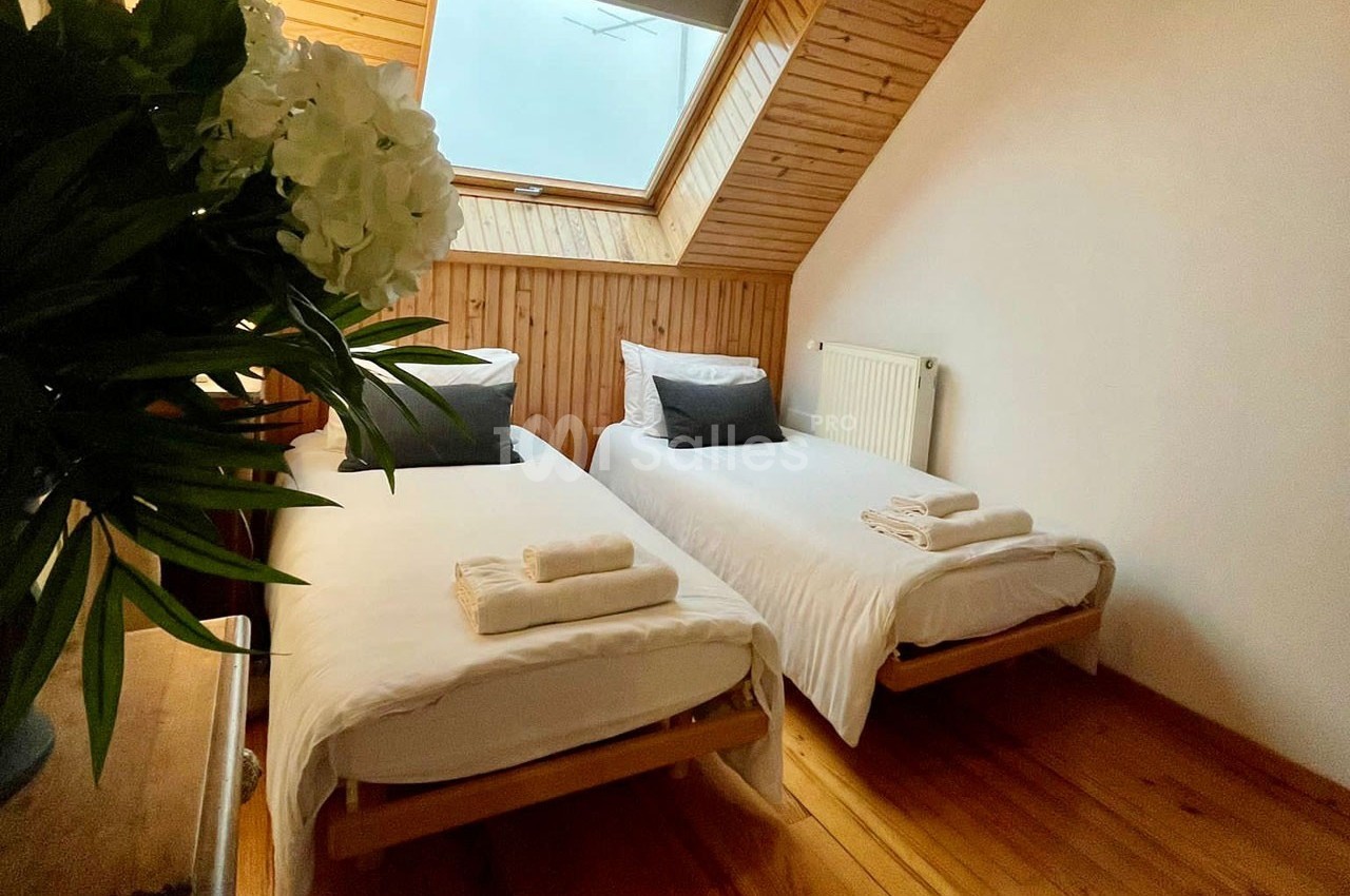 Deux lits simples avec draps blancs et coussins noirs dans une chambre mansardée avec parquet et fenêtre de toit.