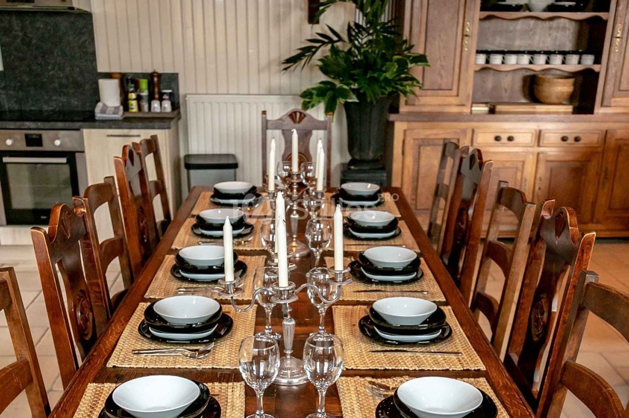 Table en bois dressée avec vaisselle, verres et chandeliers, entourée de chaises dans une salle à manger rustique.