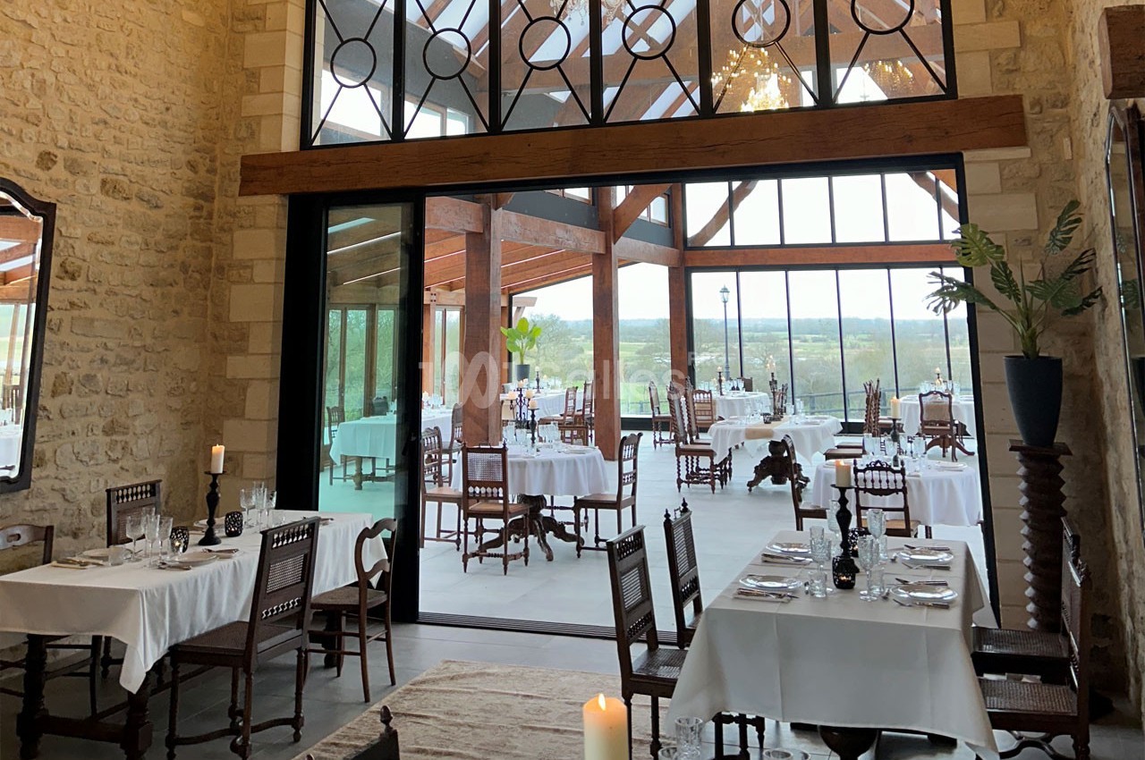 Salle de restaurant lumineuse avec murs en pierre, grandes baies vitrées et tables dressées avec nappes blanches.
