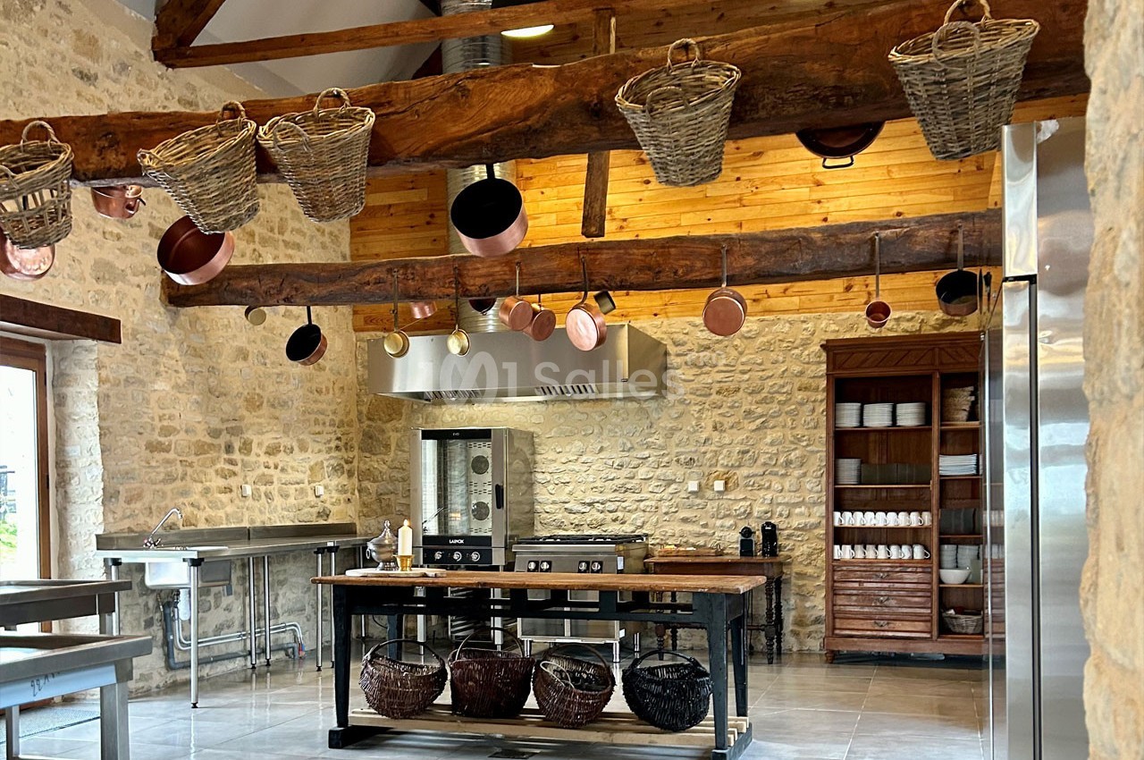 Cuisine rustique avec murs en pierre, poutres en bois, paniers suspendus et équipements modernes en inox.