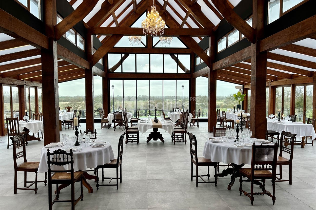 Salle de restaurant lumineuse avec de grandes baies vitrées, des tables dressées et une charpente en bois apparente.