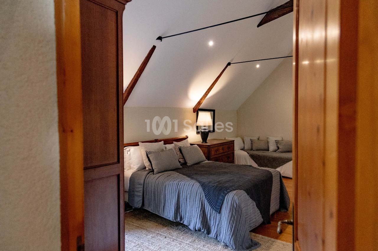 Chambre mansardée avec lit double, couvre-lit gris, lampe de chevet allumée et poutres apparentes au plafond.