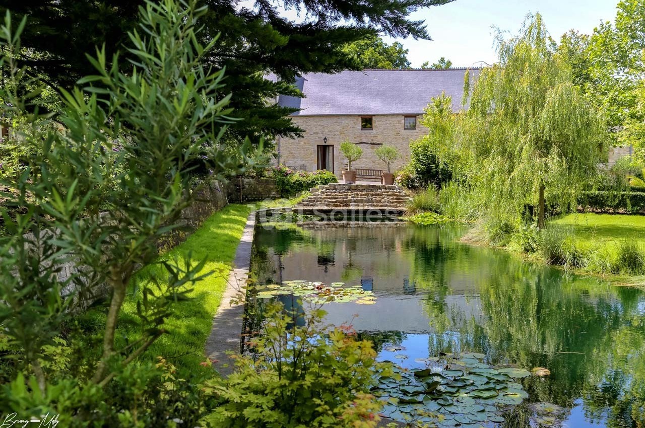 Étang bordé de végétation avec une maison en pierre et un jardin arboré en arrière-plan.