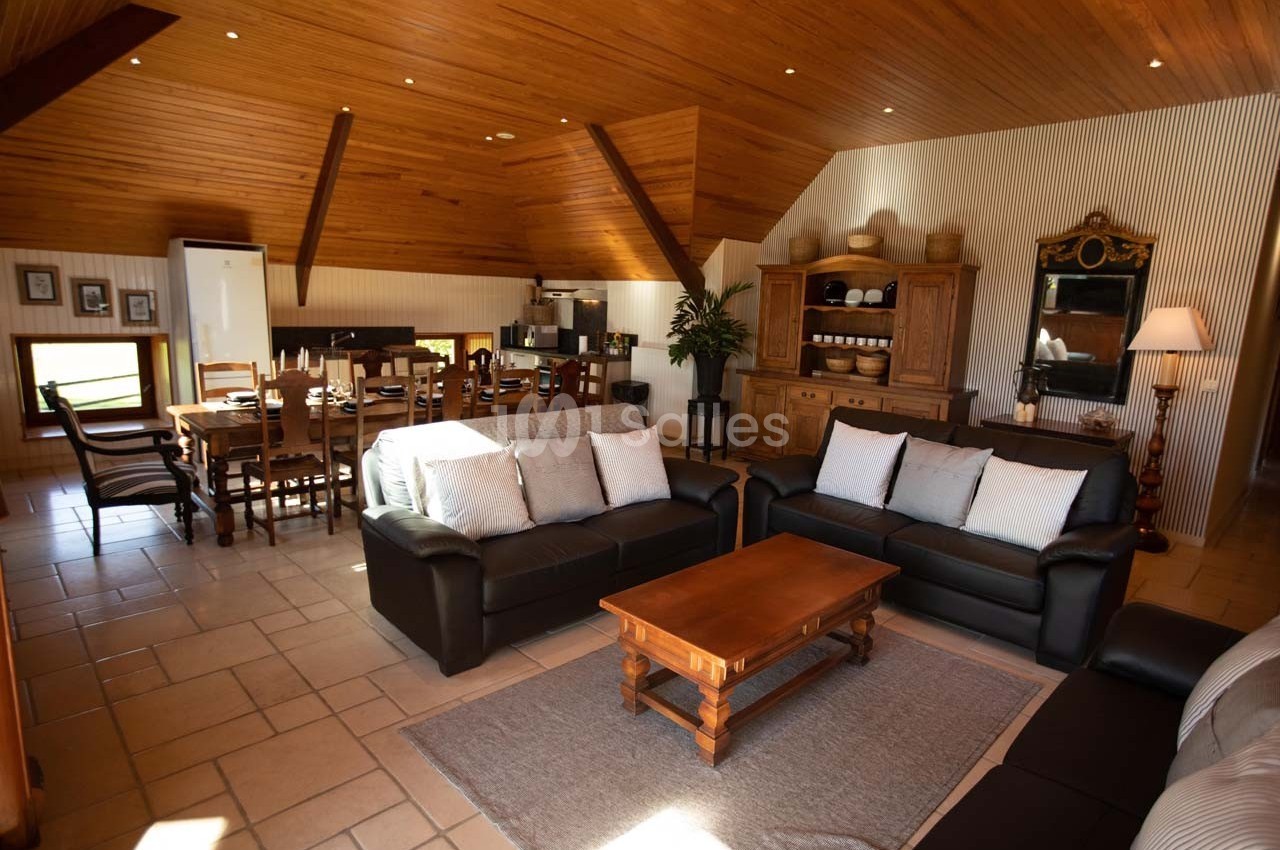 Salon spacieux avec canapés en cuir, table basse en bois, salle à manger et cuisine ouverte sous un plafond en bois.