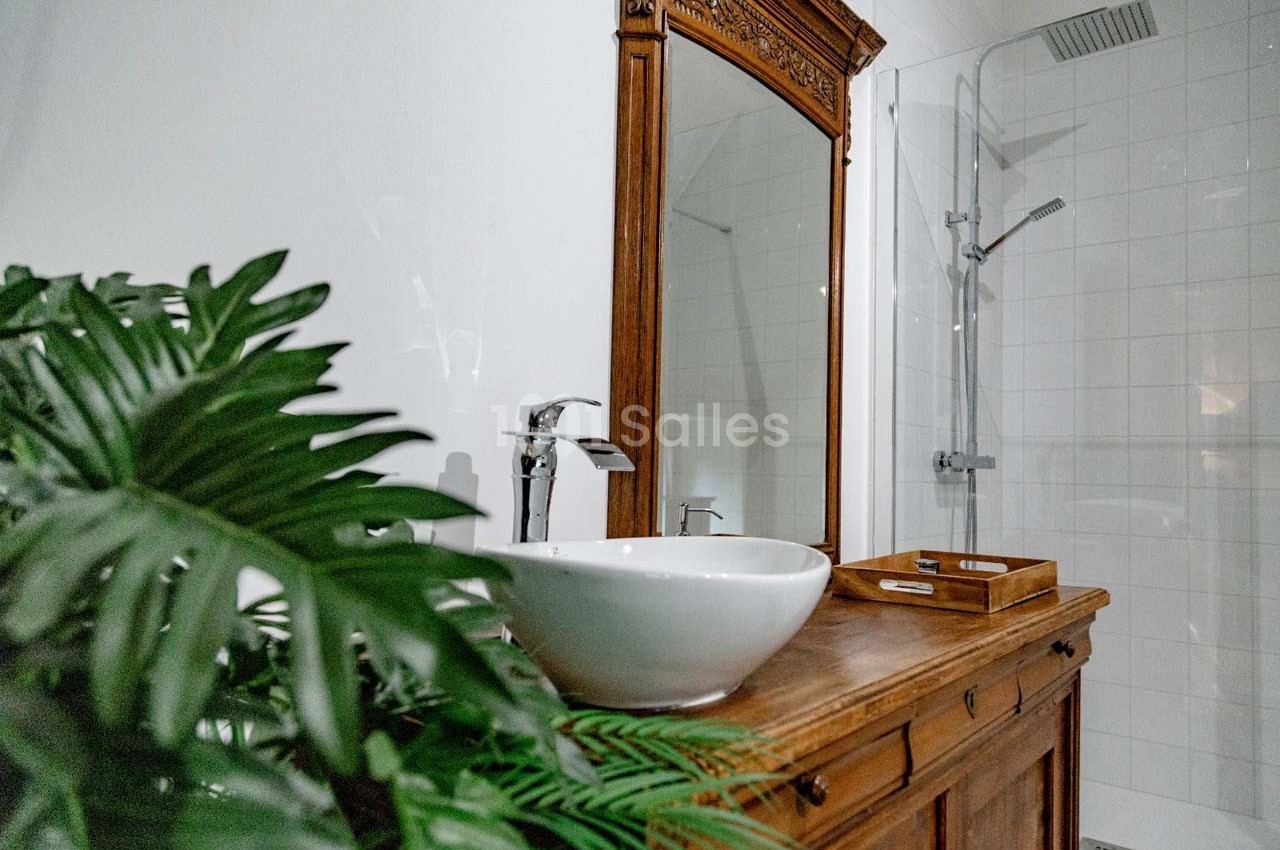 Meuble en bois avec vasque blanche, miroir encadré, plante verte et douche à l'arrière sur carrelage blanc.