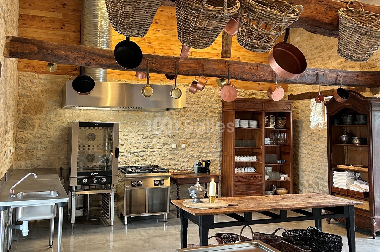 Cuisine rustique avec murs en pierre, poutres apparentes, étagères en bois, et ustensiles suspendus au plafond.
