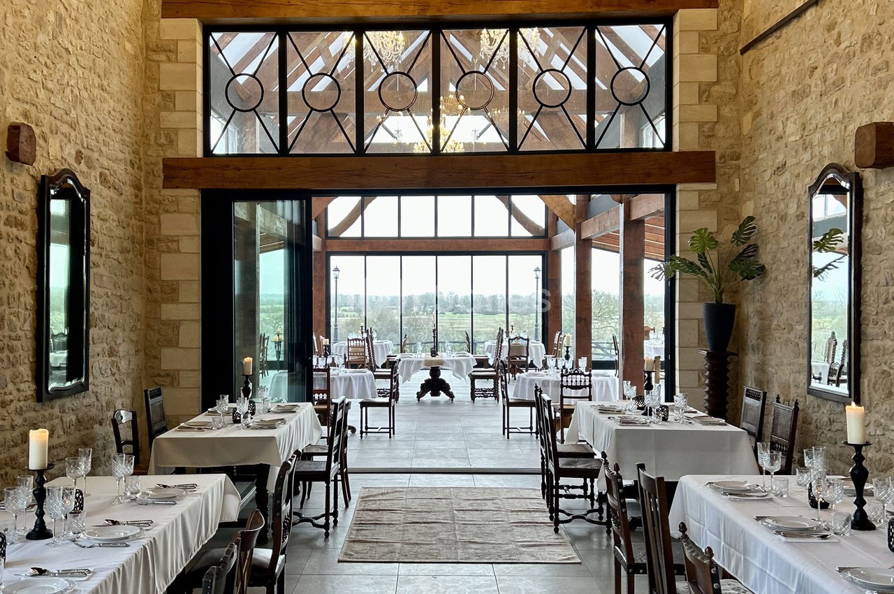 Salle de restaurant lumineuse avec murs en pierre, grandes baies vitrées et tables dressées avec nappes blanches.