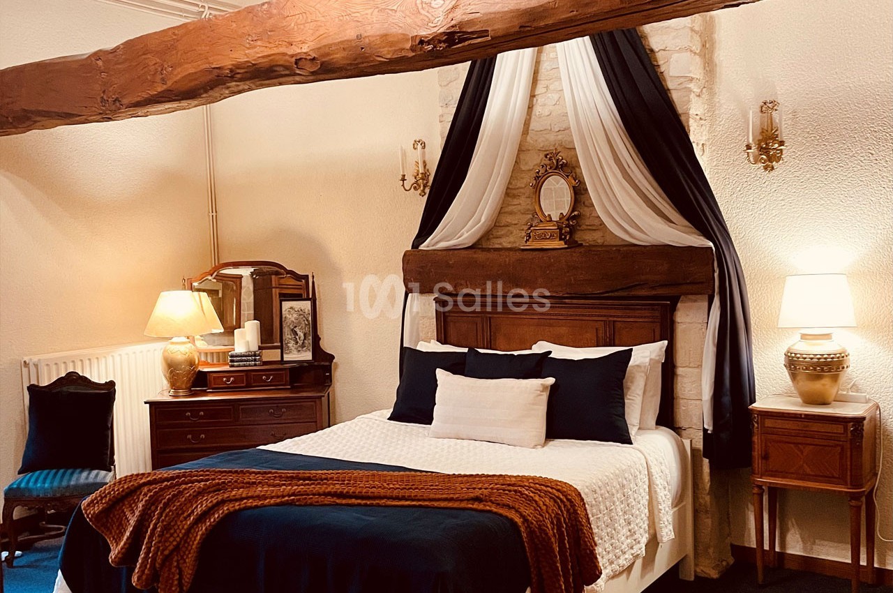 Chambre chaleureuse avec lit double, poutre apparente, draperies décoratives, meubles en bois et éclairage doux.