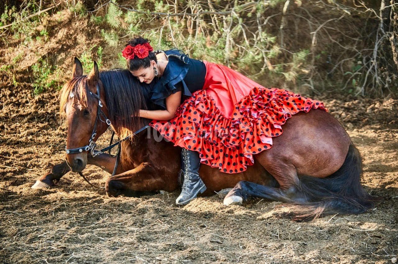 Femme en robe rouge à pois noirs allongée sur un cheval brun couché dans un environnement naturel.
