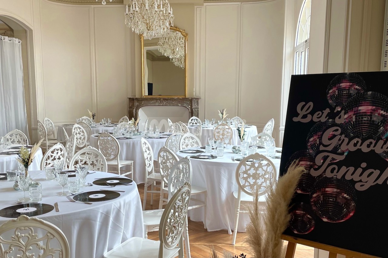 Salle élégante avec tables rondes dressées, chaises blanches décorées et panneau noir ’Let's Groove Tonight’.