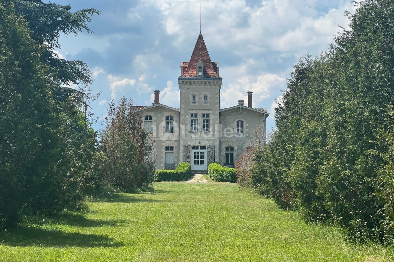 Manoir en pierre avec une tour centrale, entouré de verdure et d'arbres, sous un ciel partiellement nuageux.