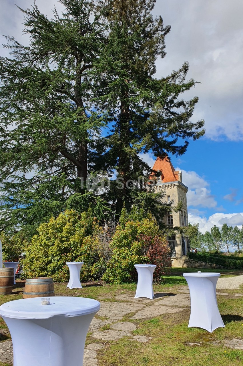 Tables hautes recouvertes de nappes blanches disposées en extérieur devant un château entouré de verdure.