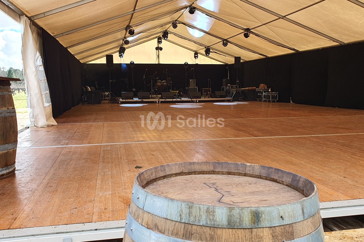 Scène vide sous un chapiteau avec parquet en bois, éclairages suspendus et tonneaux en premier plan.