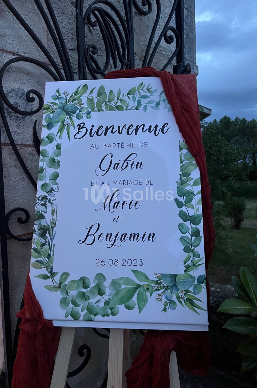 Panneau décoratif avec feuillage vert annonçant un baptême et un mariage, posé sur un chevalet en extérieur.