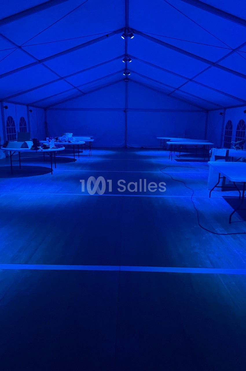 Intérieur d'une grande tente éclairée en bleu, avec des tables vides alignées sur un sol en bois.