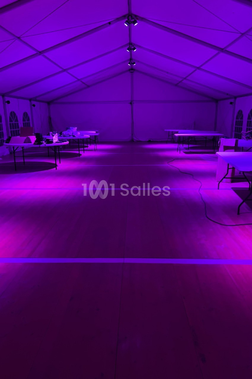 Intérieur d'une grande tente éclairée par une lumière violette, avec des tables disposées et des câbles au sol.