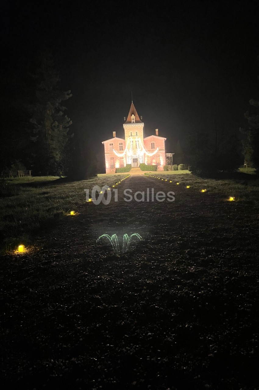 Château illuminé de nuit avec une allée bordée de petites lumières au sol, entouré de végétation sombre.
