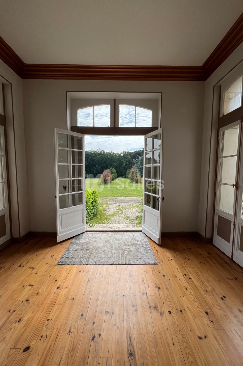 Vue depuis une pièce lumineuse avec parquet, montrant des portes-fenêtres ouvertes sur un jardin verdoyant.