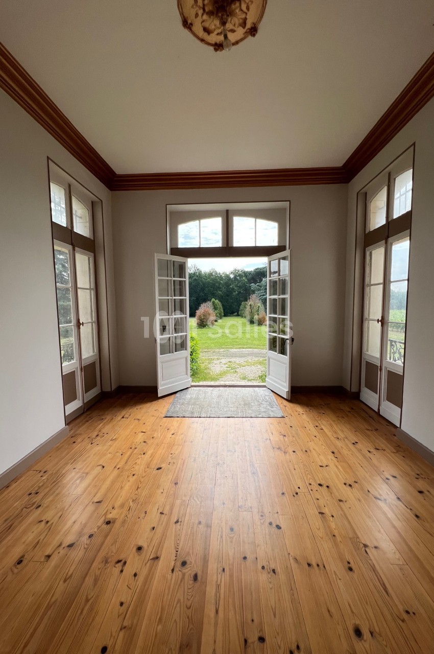 Salle lumineuse avec parquet en bois, portes-fenêtres ouvertes donnant sur un jardin verdoyant et un ciel partiellement…