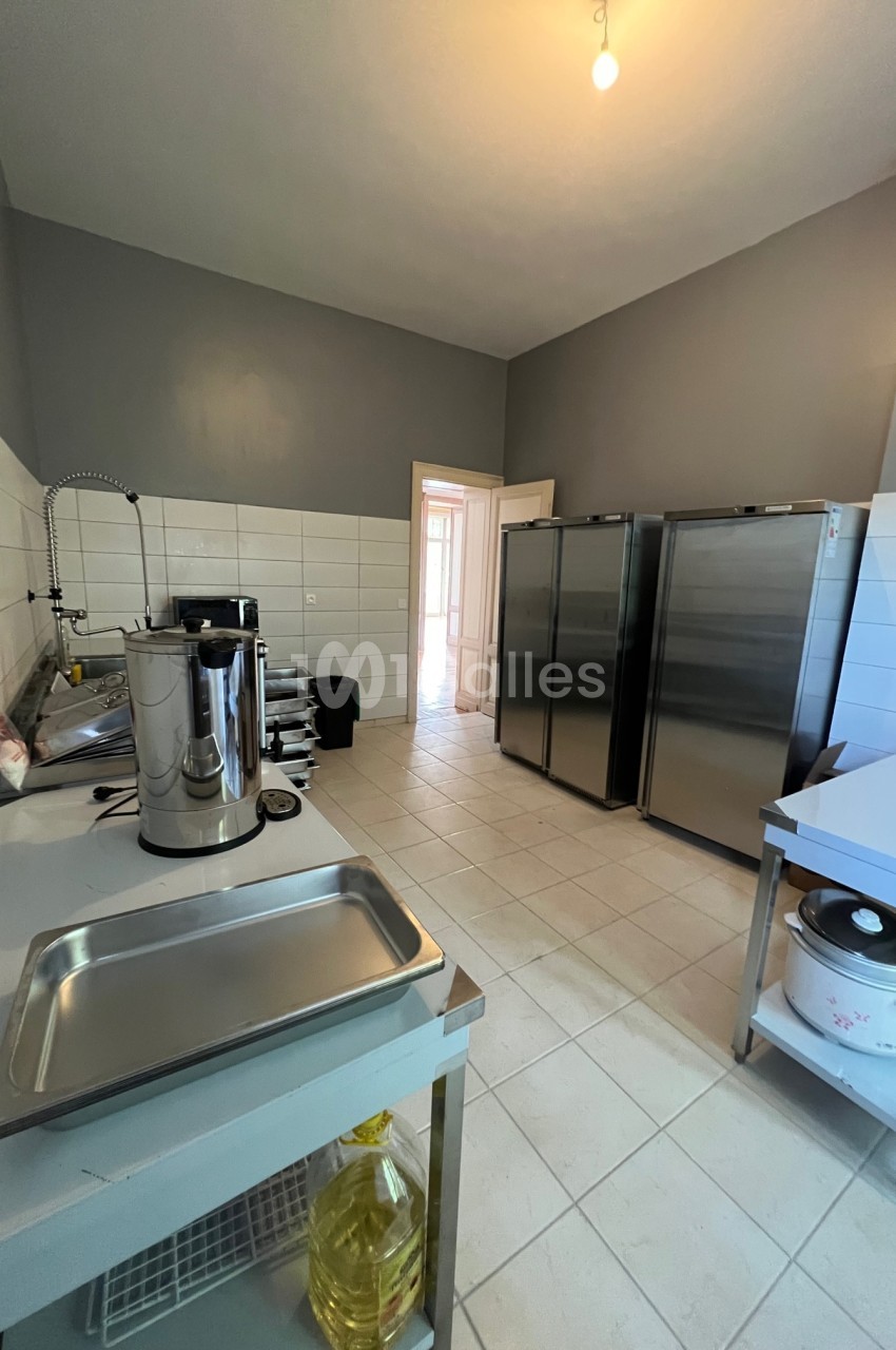 Cuisine professionnelle avec équipements en inox, réfrigérateurs, plans de travail et carrelage clair au sol et aux murs.