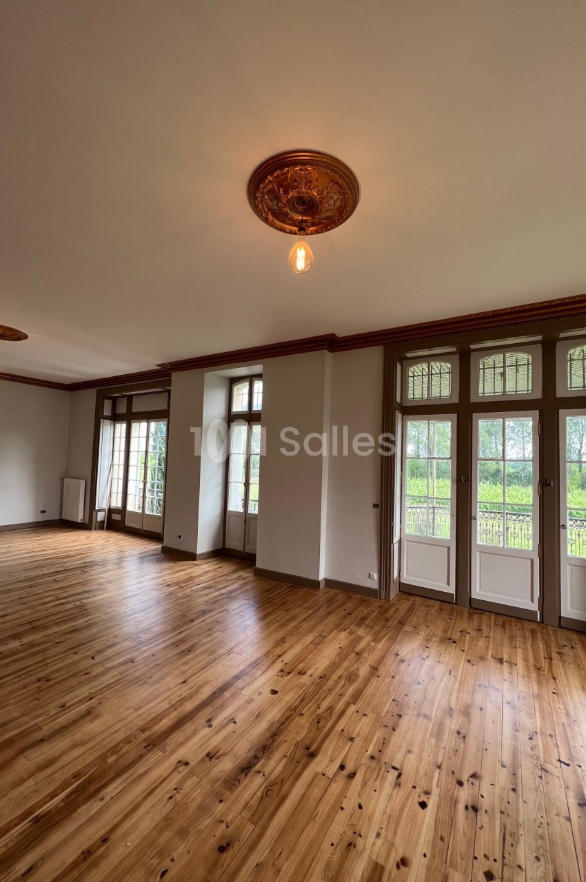 Salle lumineuse avec parquet en bois, moulures au plafond et grandes fenêtres donnant sur un espace vert.