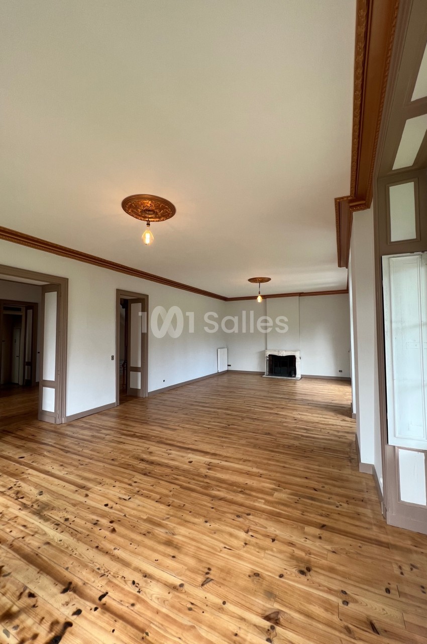 Grande pièce lumineuse avec parquet en bois, moulures au plafond, cheminée et grandes fenêtres donnant sur l'extérieur.