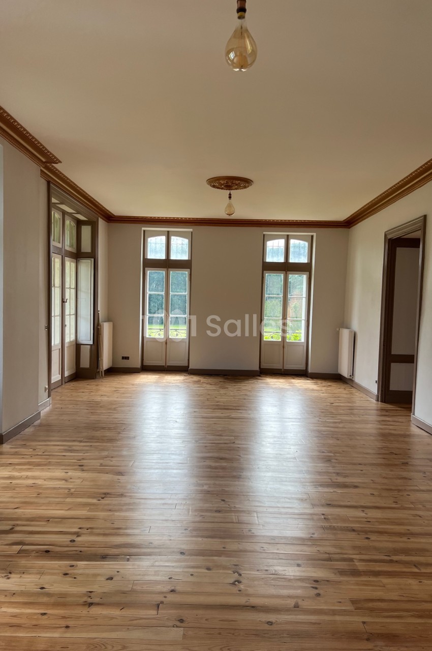 Salle lumineuse avec parquet en bois, grandes fenêtres à double battant et moulures au plafond.