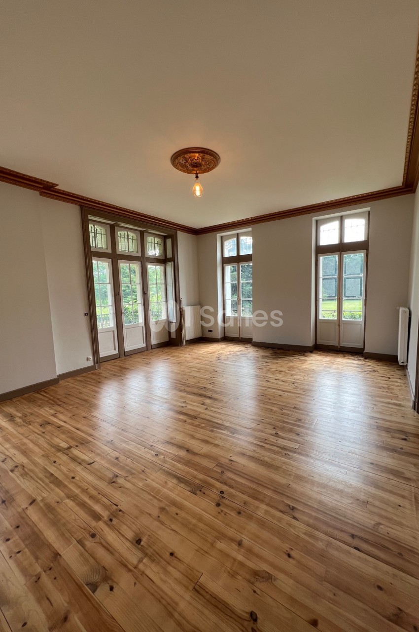 Salle lumineuse avec parquet en bois, grandes fenêtres donnant sur un jardin et moulures au plafond.