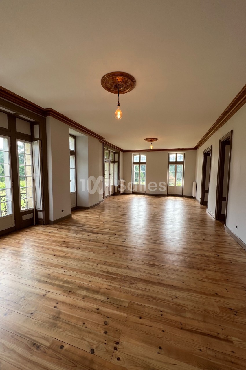 Salle lumineuse avec parquet en bois clair, grandes fenêtres et moulures au plafond, donnant sur un espace verdoyant.