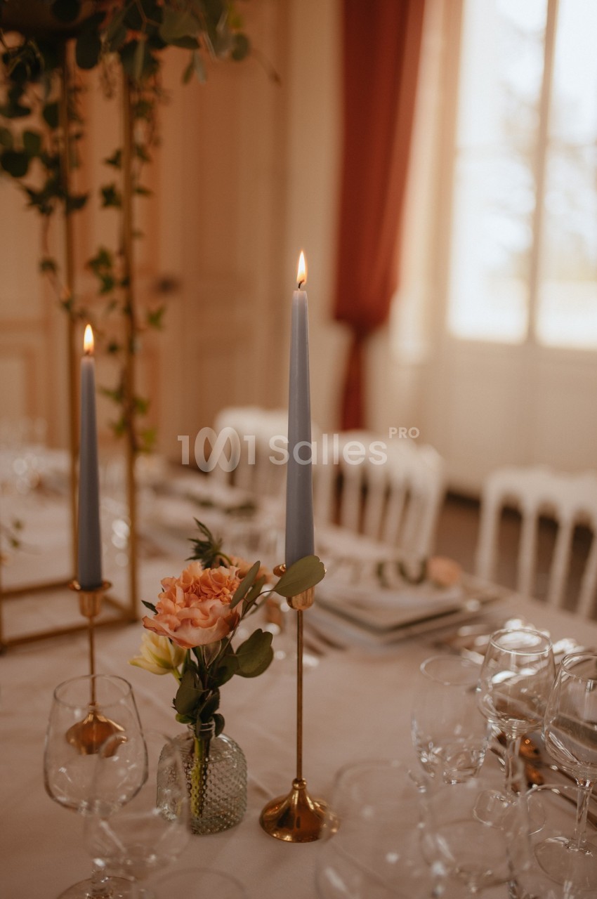 Bougies allumées et fleurs dans un vase décorent une table élégamment dressée dans une pièce lumineuse.