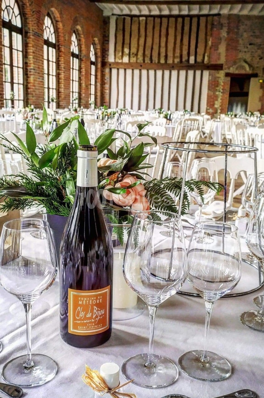 Salle de réception élégante avec tables dressées, centre de table floral et bouteille de vin au premier plan.